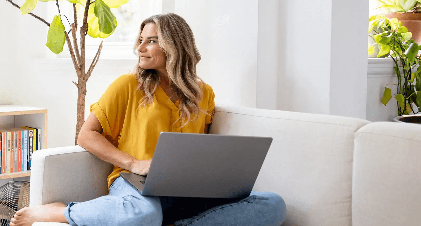 A woman sits cross-legged on a sofa with a laptop, surrounded by bookshelves and plants.